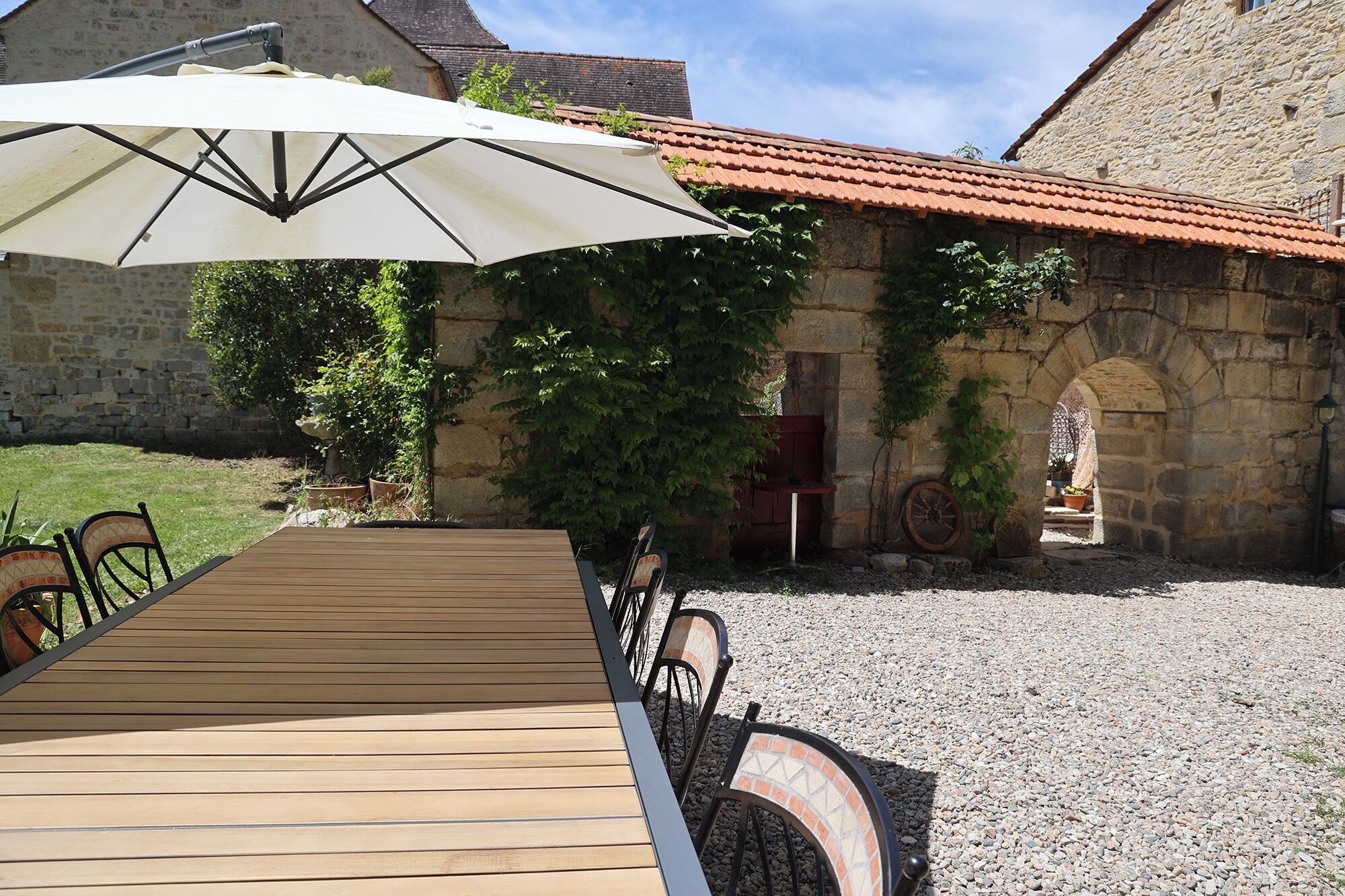 A wooden table with chairs and a white umbrella in a courtyard with stone walls and a tiled roof.