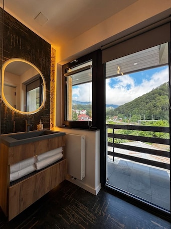 A modern bathroom with a wooden vanity, a large mirror, and a view of a mountainous landscape through a sliding glass door.