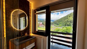 Ein Badezimmer mit einem großen Spiegel, einer Holzkommode und Blick auf eine grüne Landschaft durch ein großes Fenster.