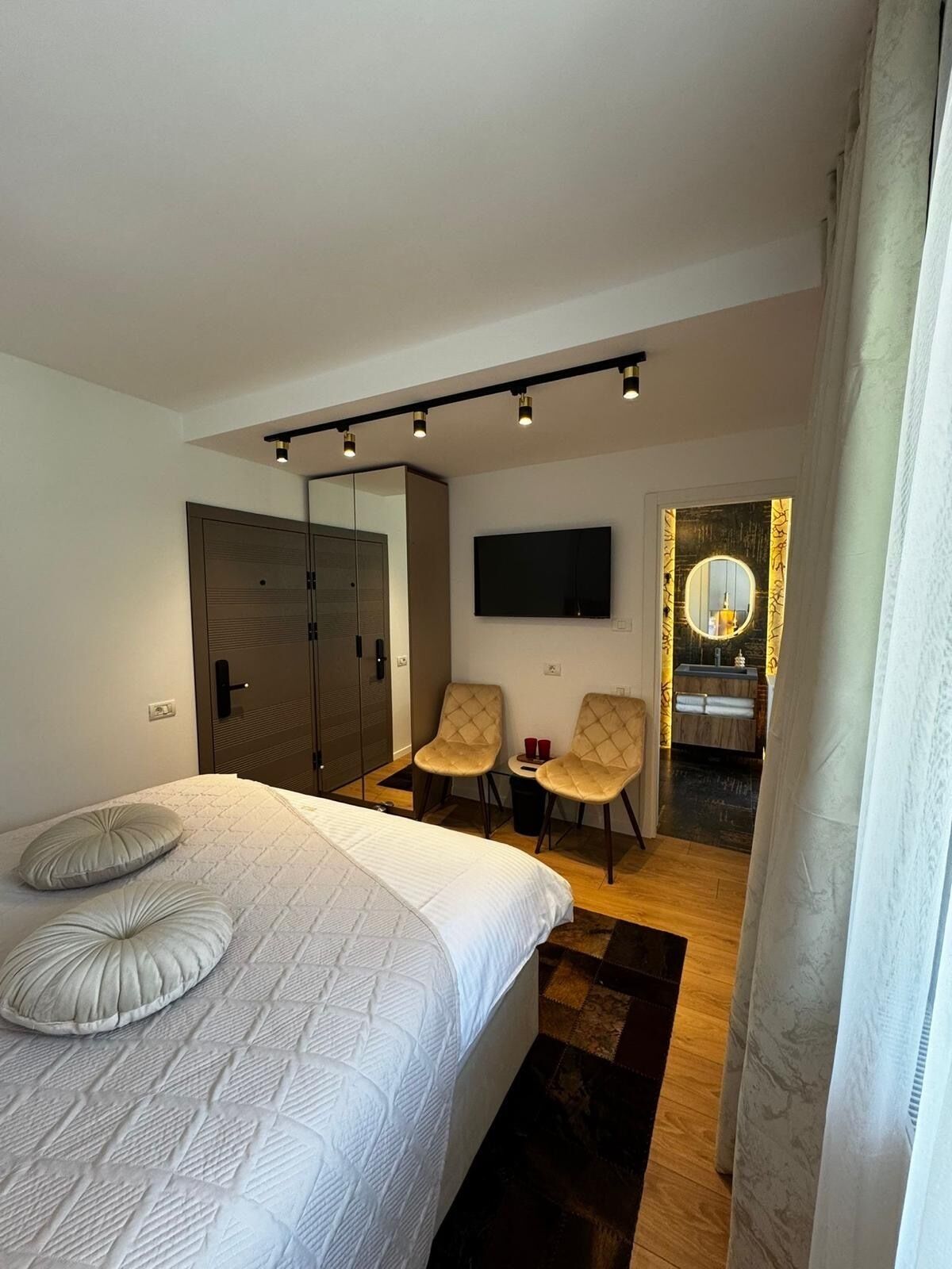 Ein modernes Hotelzimmer mit einem großen Bett, zwei Sesseln, einem Fernseher, einem Spiegel und einem kleinen Badezimmer.