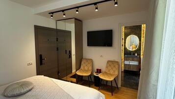 Ein modernes Hotelzimmer mit einem großen Bett, zwei Sesseln, einem Fernseher, einem Spiegel und einem kleinen Badezimmer.
