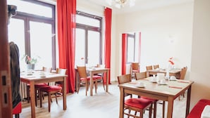 A dining area with wooden tables and red chairs, large windows with red curtains, and a white wall with a small plant. - Hotel Villa Kisseleff (Bad Homburg v.d. Hoehe)