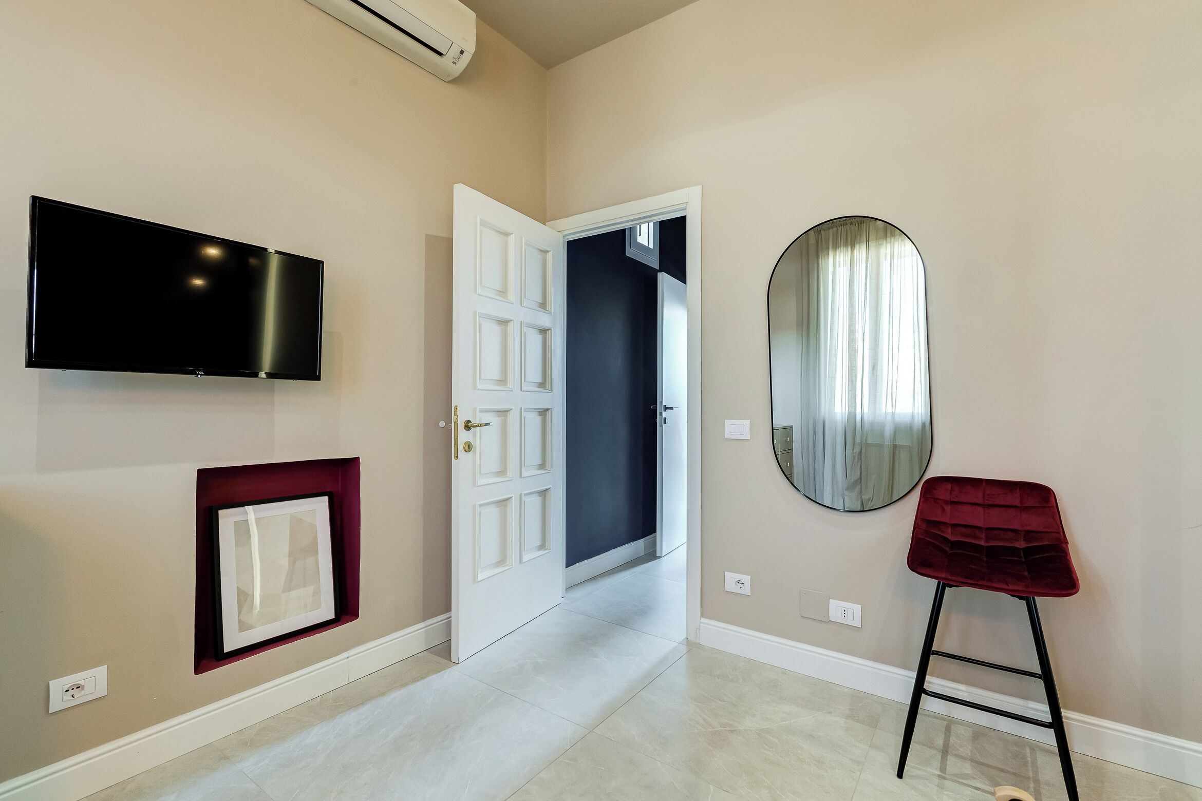 A hallway with a flat-screen TV, a door, a mirror, and a red stool.