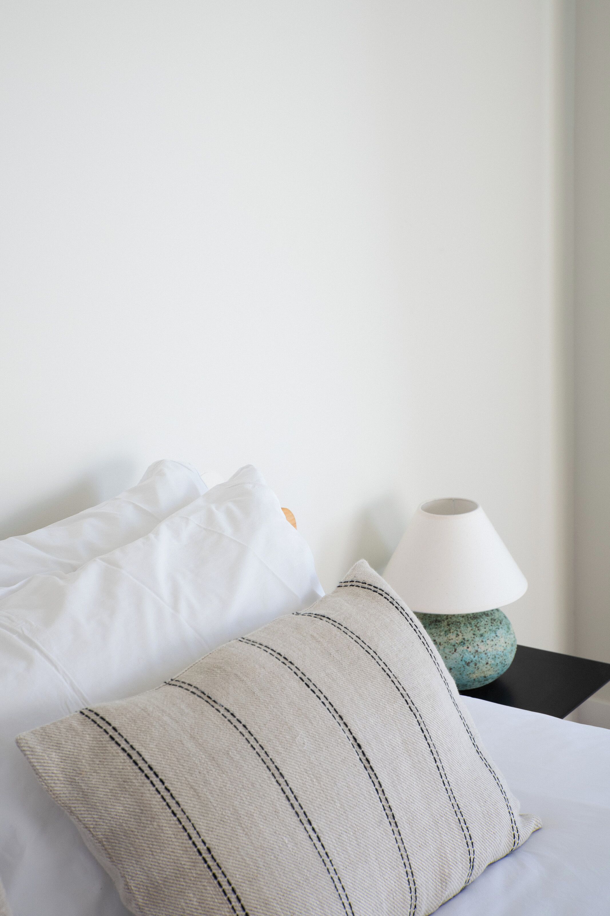 Un lit bien fait, avec des oreillers blancs et rayés, une lampe blanche posée sur une table de chevet noire et un mur sobre.