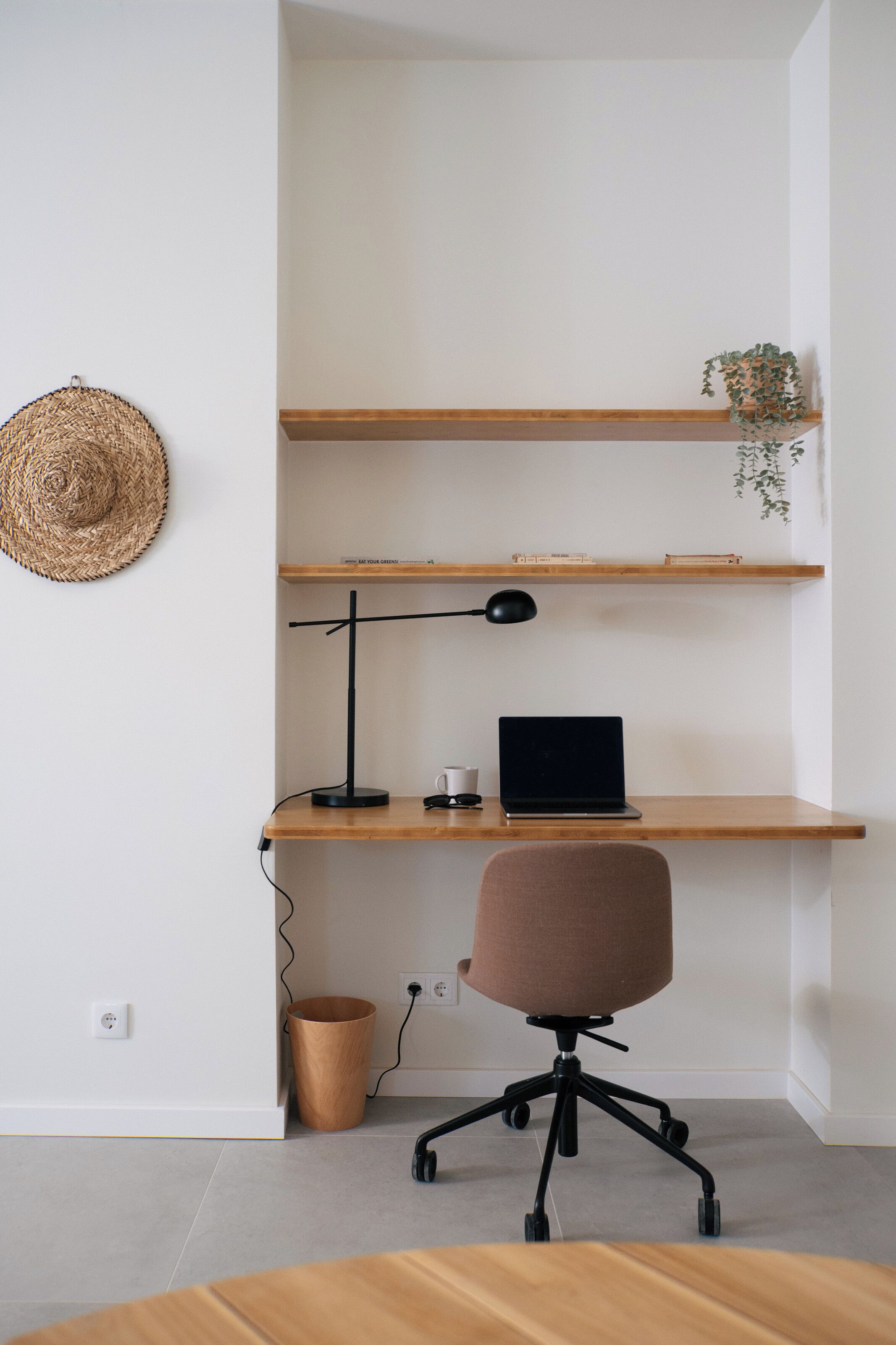 A workspace with a laptop, desk lamp, and office chair, with shelves above and a straw hat on the wall.