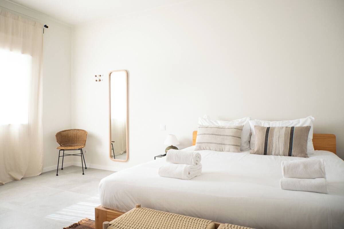 A neatly arranged bedroom with a large bed, a chair, a mirror, and a nightstand with a lamp.