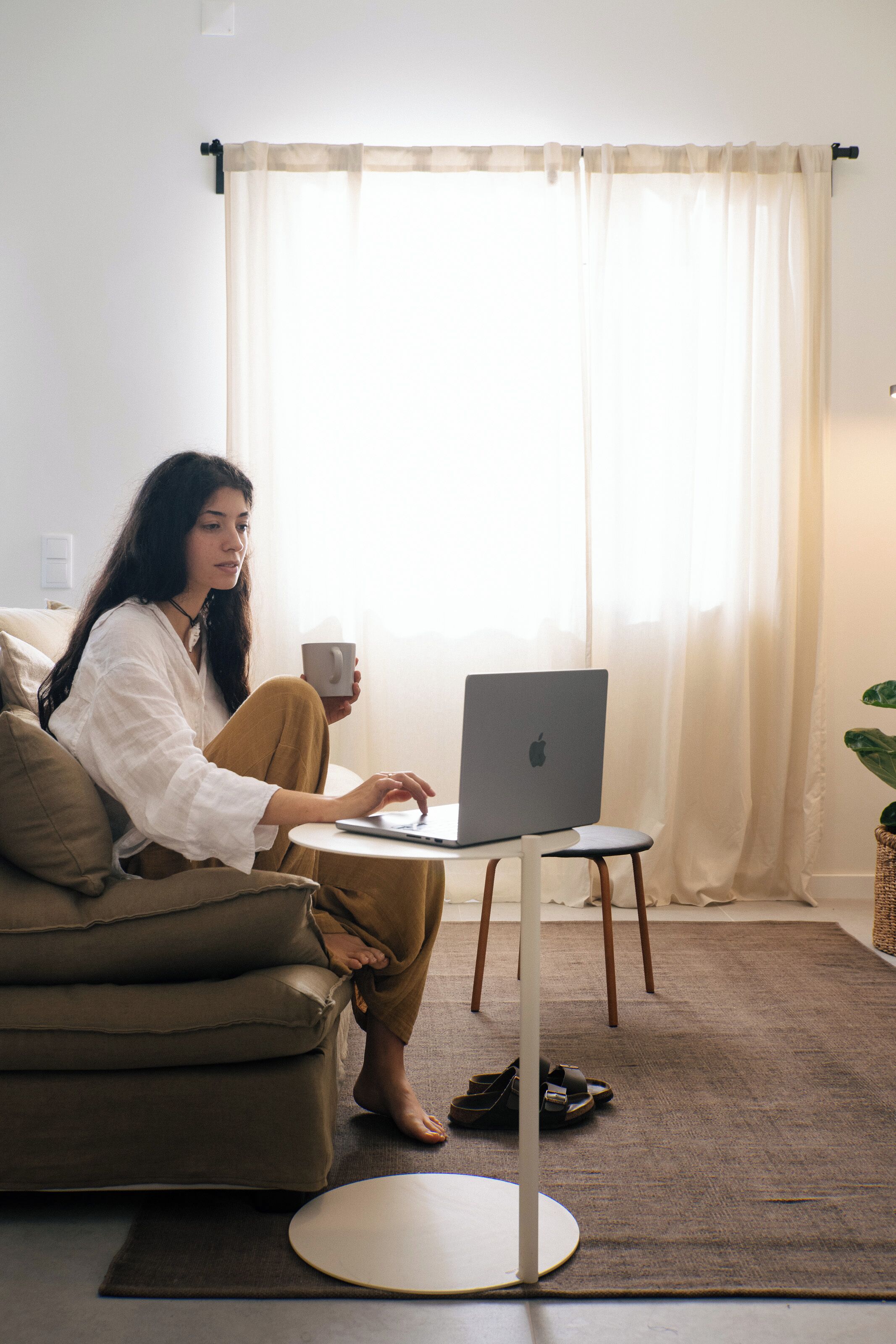On peut voir une personne assise sur un canapé, un ordinateur portable sur les genoux, tenant une tasse et ayant une plante à proximité.