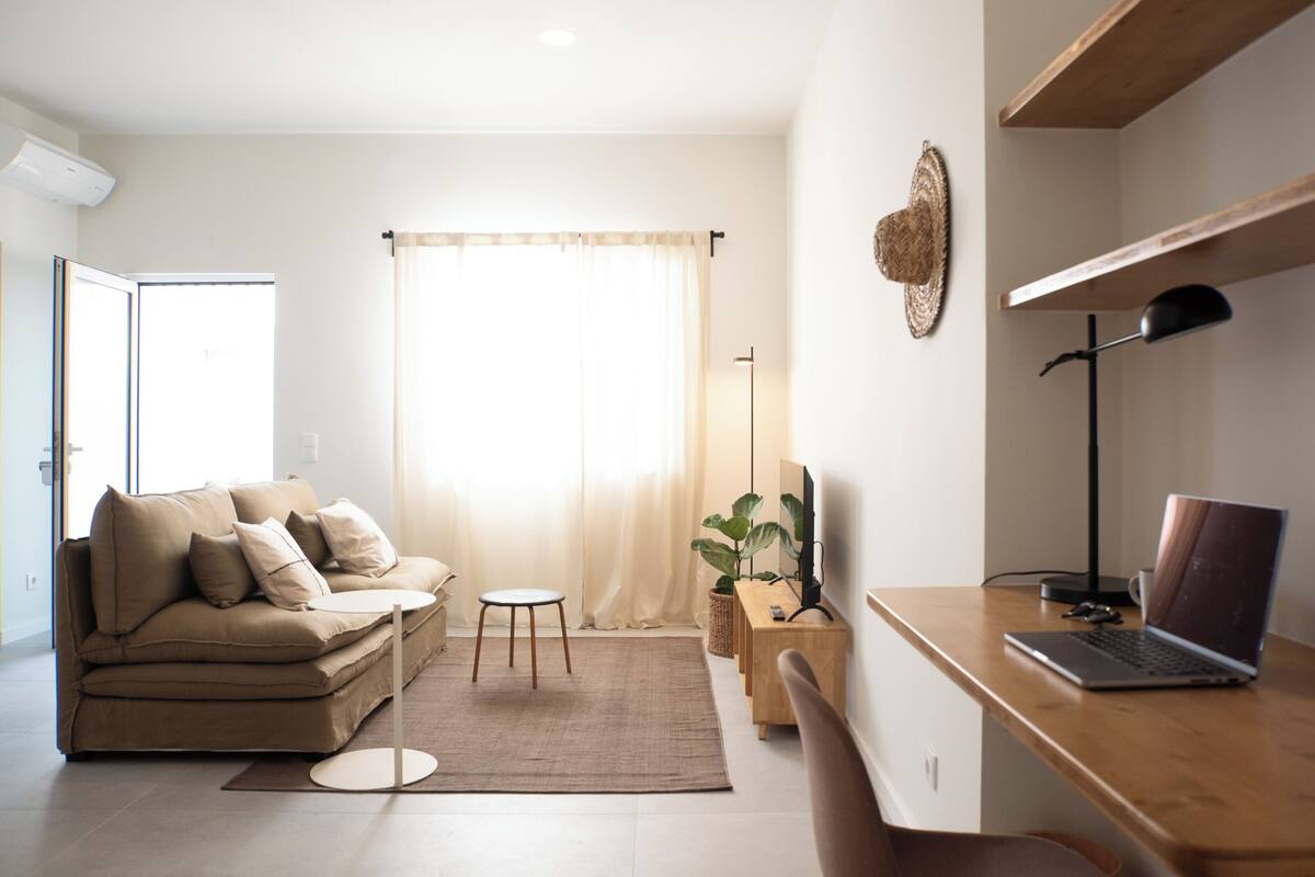 A modern living room with a sofa, a coffee table, a desk with a laptop, a floor lamp, and a large window with curtains.