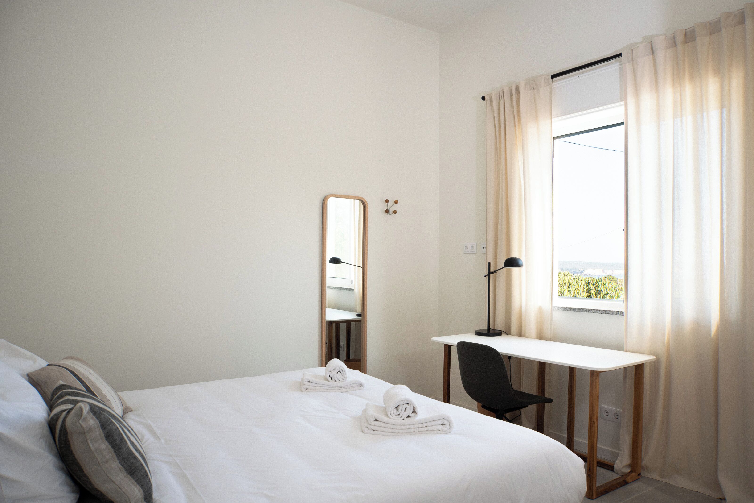 Une chambre d’hôtel avec un lit, un bureau, une chaise, un miroir et une fenêtre avec des rideaux.