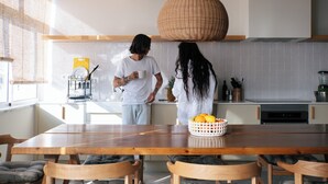 Une cuisine moderne avec une table à manger en bois, deux personnes en train de discuter et un panier de fruits sur la table.