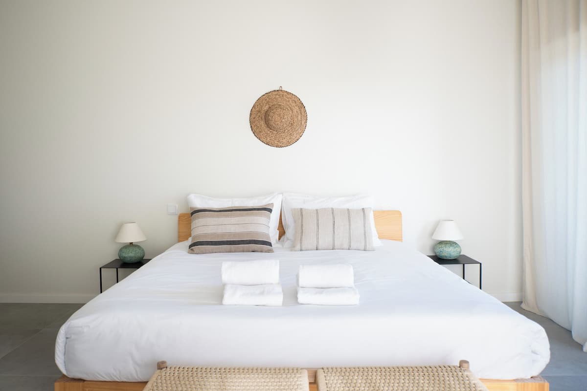 A neatly made bed with white linens, two pillows, and two folded towels. There is a woven wall hanging above the bed, two bedside tables with lamps, and a woven mat in front of the bed.