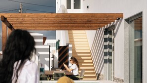 Un espace piscine avec une terrasse en bois, une personne assise au bord, et un bâtiment à la façade blanche surmonté d’une pergola en bois.