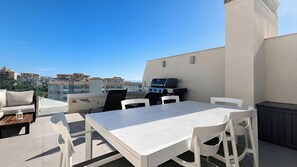 Eine Dachterrasse mit einem weißen Tisch und Stühlen, einem Grill und Blick auf die Stadtlandschaft.