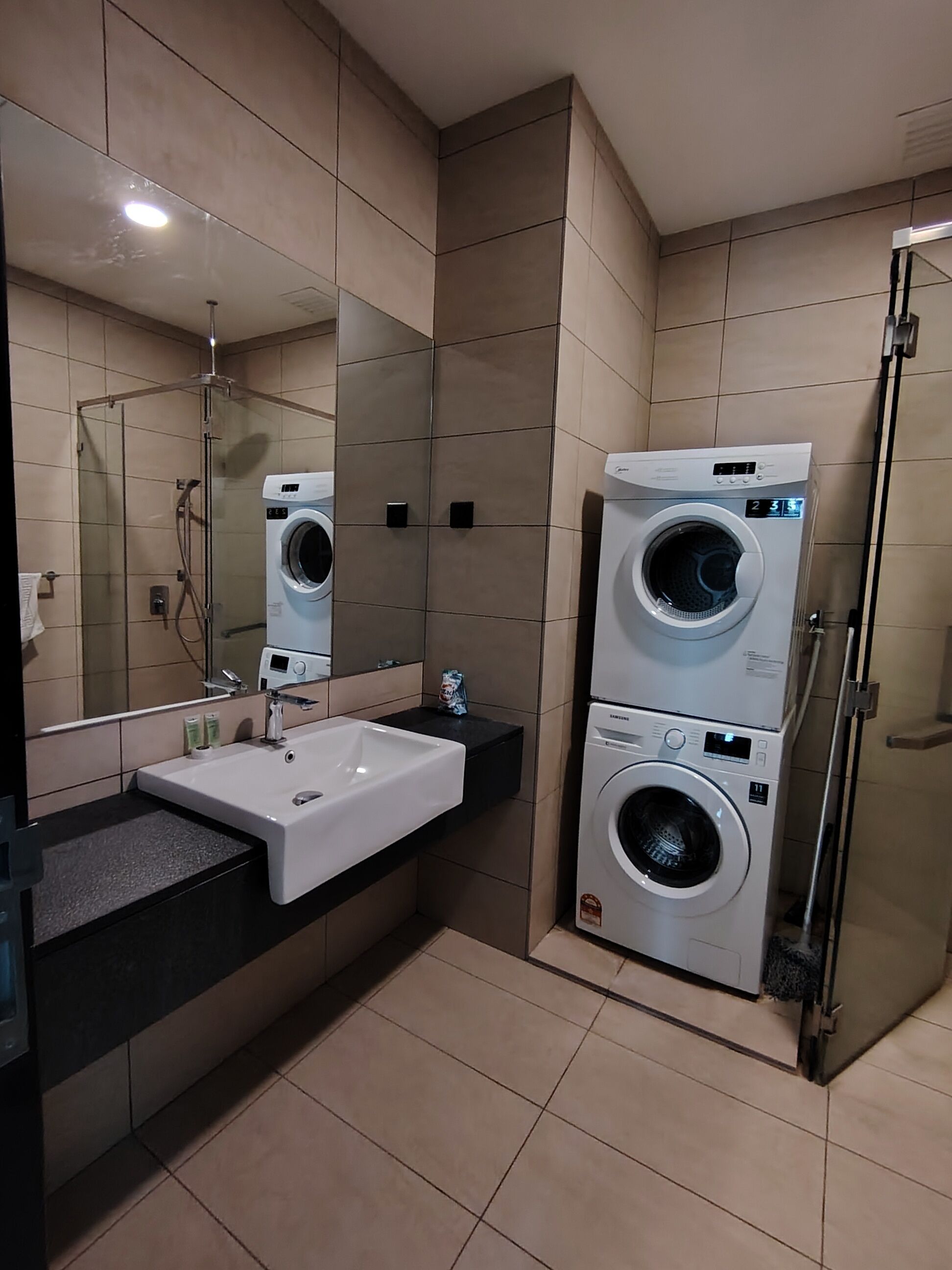 A modern bathroom with a large mirror, a sink, two stacked washing machines, and a shower area.