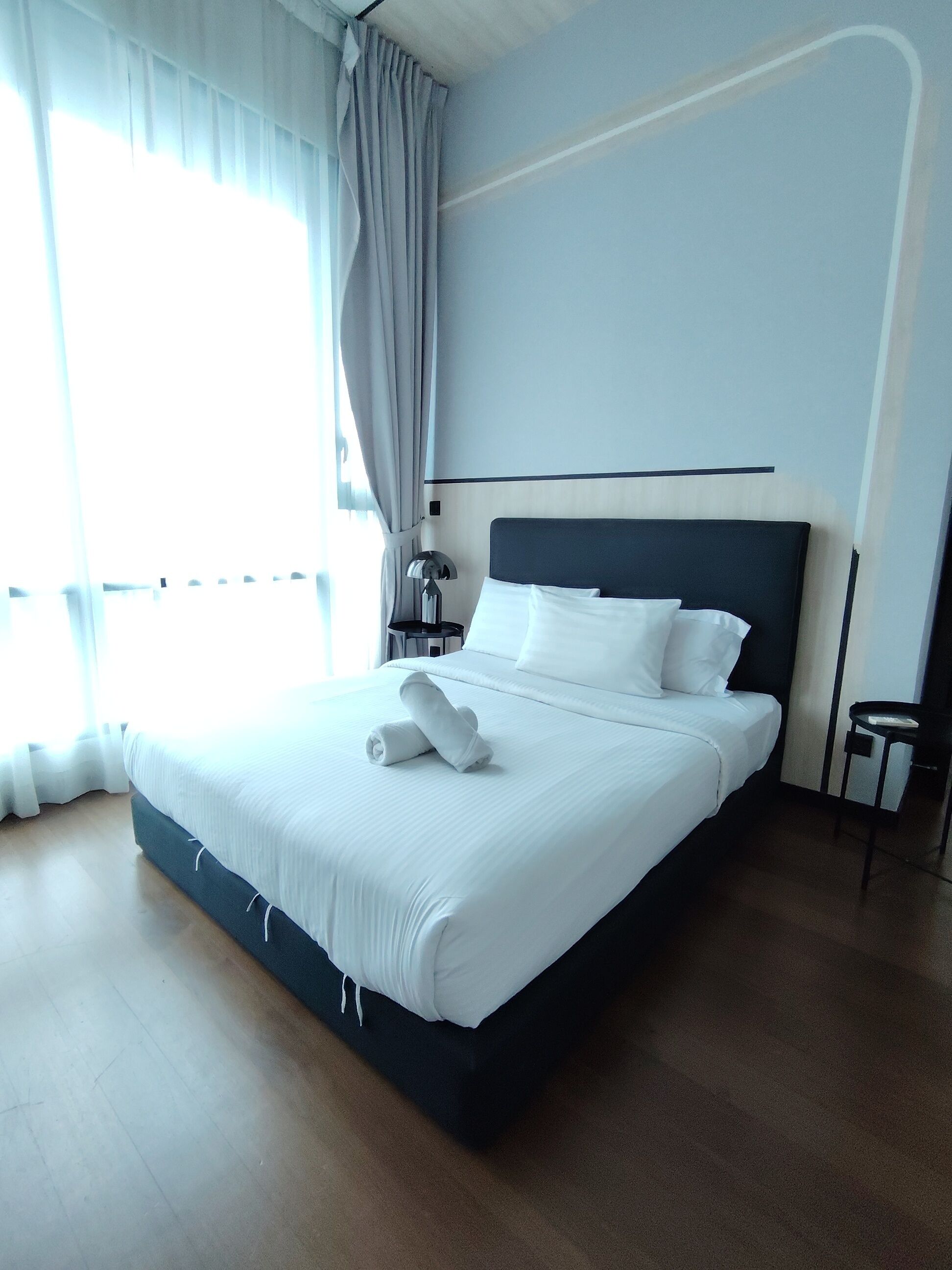 Een netjes opgemaakt bed met witte lakens en een opgevouwen handdoek, een nachtkastje met een lamp, en uitzicht op een raam met gordijnen.