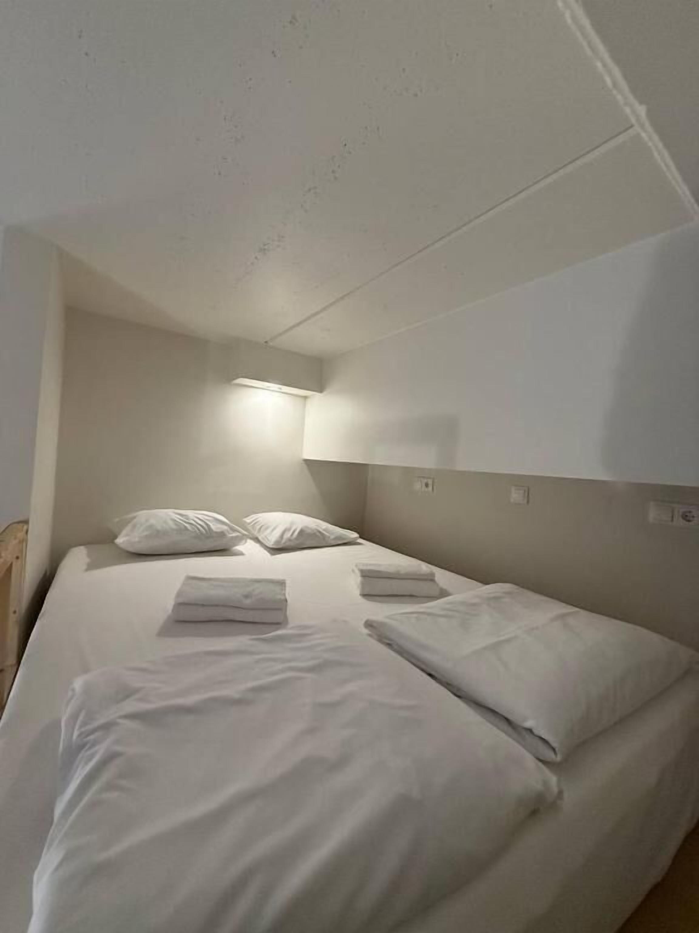 Una cama individual con ropa de cama y almohadas blancas, una mesita de noche y una lámpara de pared.