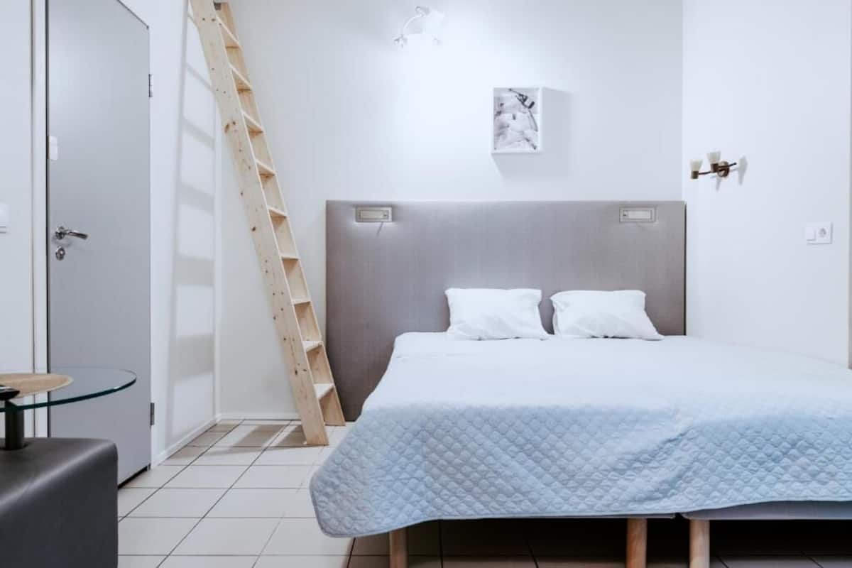 A bedroom with a bed, a ladder, a small table, and a wall-mounted light fixture.