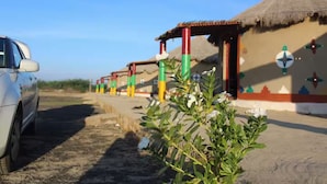 A row of thatched-roof huts with colorful pillars, a parked car, and a clear sky. - Rann Roshni Homestay – Family Friendly Stay in Kutch 6 (Bhirandiara)