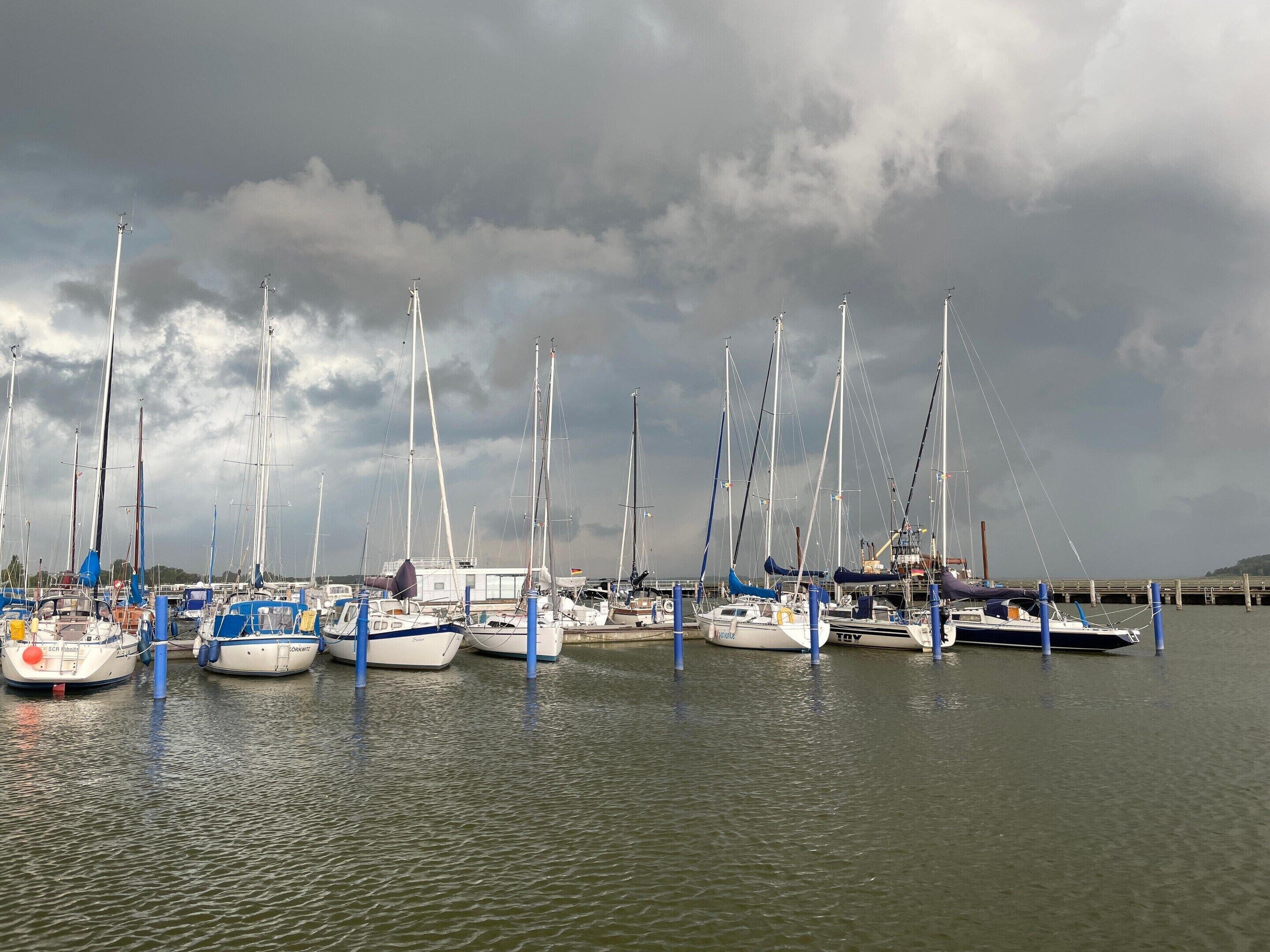 A marina with multiple sailboats docked, some with blue fenders, under a cloudy sky.