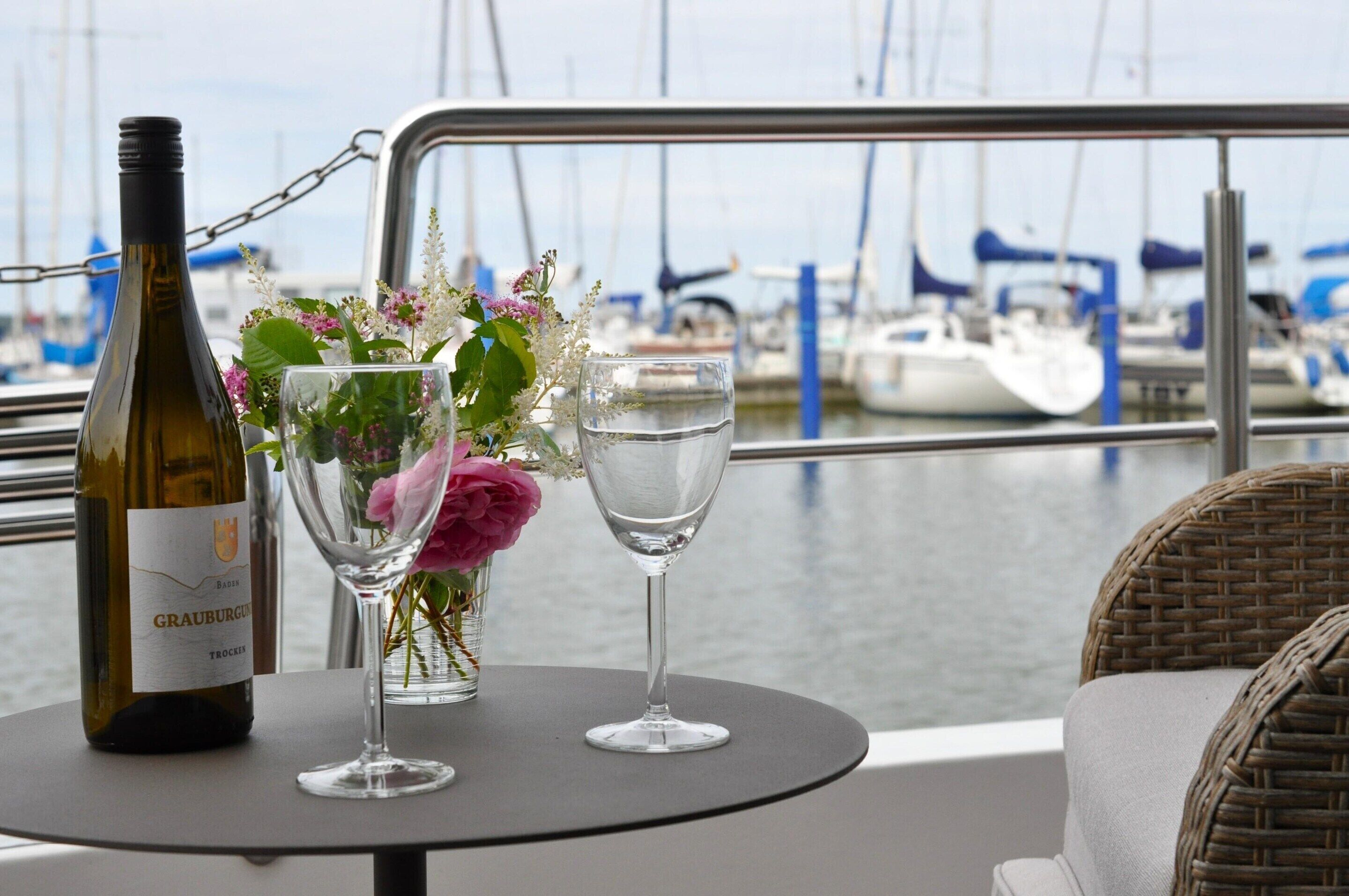 A bottle of wine, a glass with flowers, and a glass of water on a table with a view of boats.