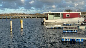 A stationary boat docked at a pier with a small cabin structure, surrounded by calm water and a cloudy sky. - Hausboot Paula mit Reisekajak in Ribnitz-damgarten (Ribnitz-Damgarten)