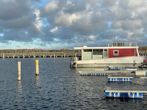 A stationary boat docked at a pier with a small cabin structure, surrounded by calm water and a cloudy sky. - Hausboot Paula mit Reisekajak in Ribnitz-damgarten (Ribnitz-Damgarten)