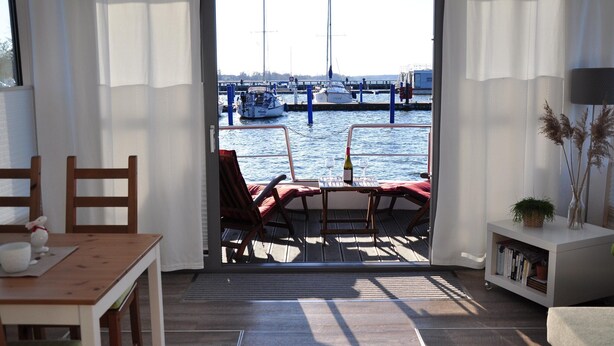 A room with a view of a marina, a wooden table, a red chair, a bookshelf, and a small plant. - Hausboot Paula mit Reisekajak in Ribnitz-damgarten (Ribnitz-Damgarten)