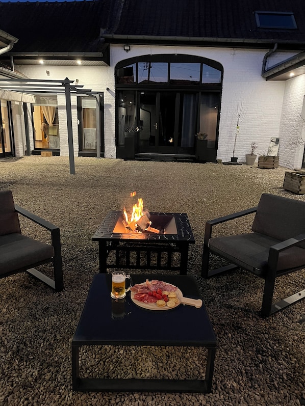 Un espace extérieur aménagé avec un foyer, une table sur laquelle se trouve une assiette de nourriture et un pichet de bière, ainsi que deux chaises.