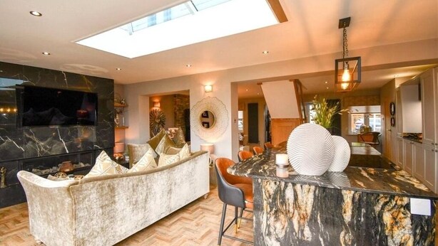 Ein modernes Wohnzimmer mit Marmor-Kamin, einer Couch mit Kissen, einem Essbereich mit Marmorinsel und einem großen Fenster, das für natürliches Licht sorgt.