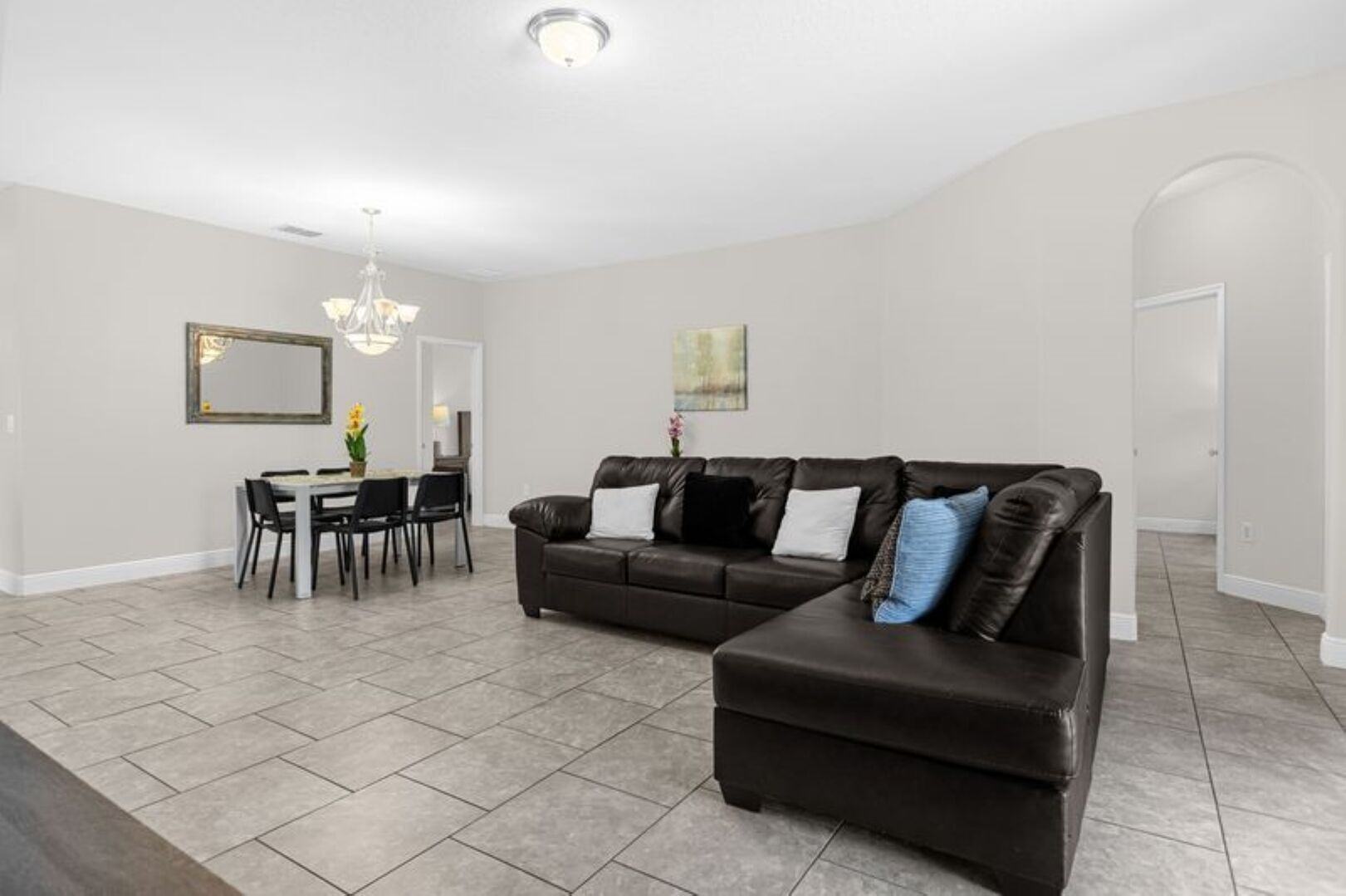 A living room with a black leather sectional sofa, a dining table with chairs, and a chandelier.