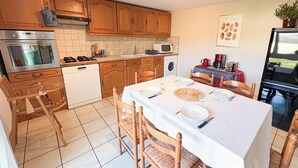 A kitchen with wooden cabinets, a dishwasher, a stove, a microwave, and a dining table set with plates and glasses. - Quiet country & family gîte 20min from Caen (Cintheaux)