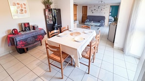 A dining area with a table set for four, chairs, a refrigerator, and a kitchen visible in the background. - Quiet country & family gîte 20min from Caen (Cintheaux)