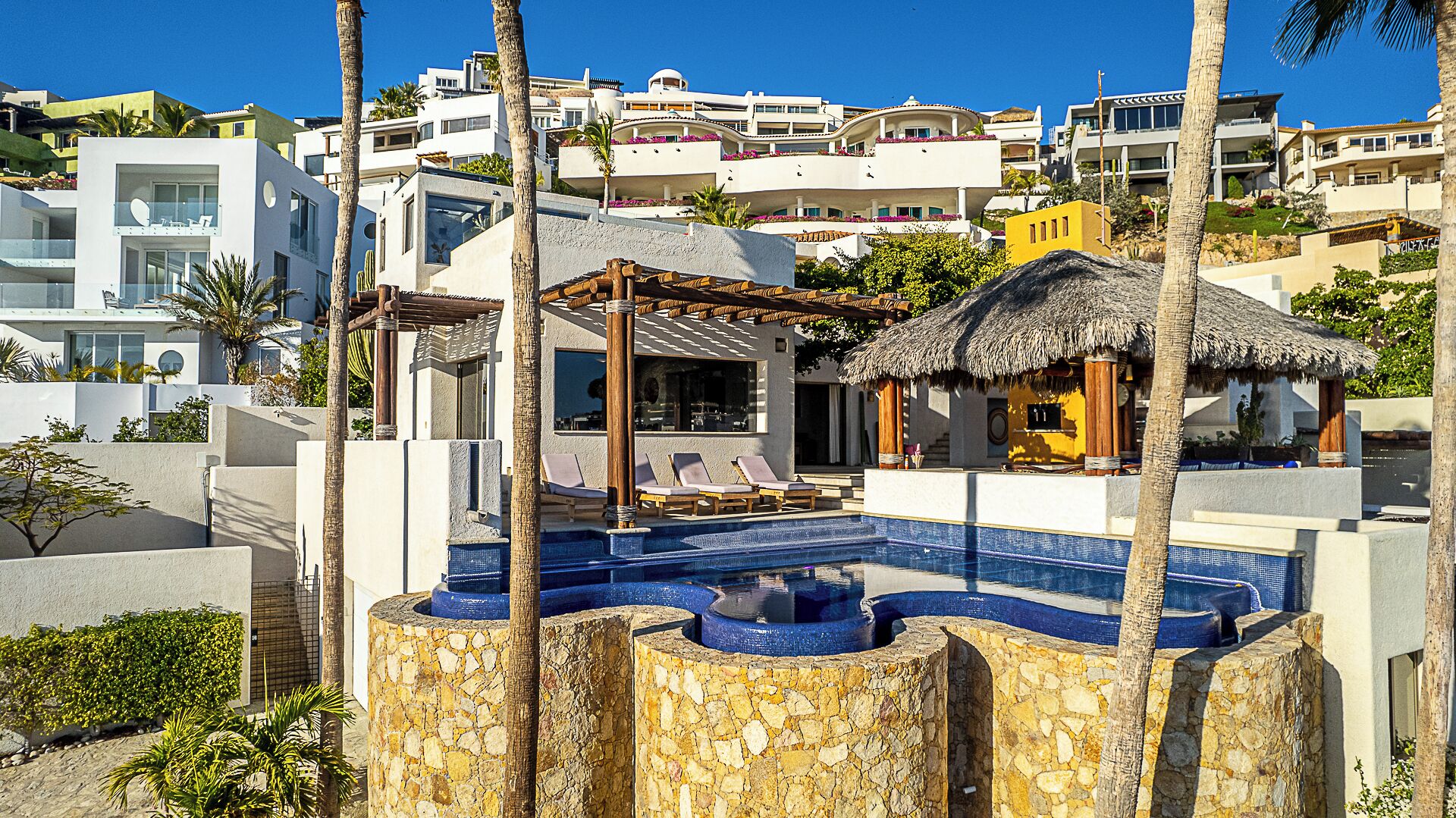 Un resort moderno con zona de piscina, que incluye un jacuzzi y sillas de descanso, rodeado de palmeras y edificios de estilo mediterráneo.