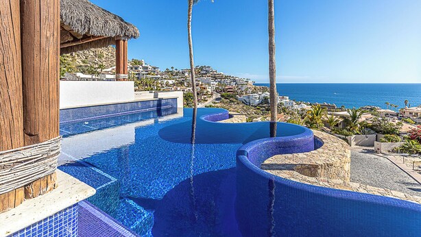 A pool with a curved design, a thatched-roof structure, and a view of a coastal town. - Casa Dos Hermanas by Wynwood House (Cabo San Lucas)