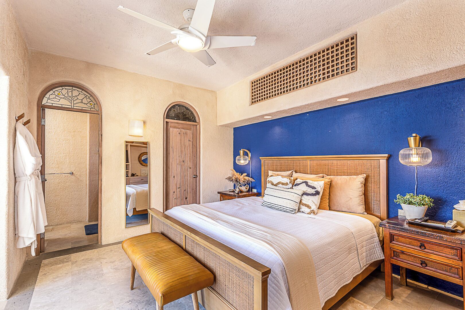 A bedroom with a large bed, a wooden bench, a ceiling fan, and a nightstand with a lamp.