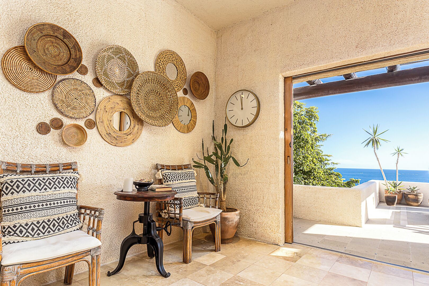 Un intérieur chaleureux, agrémenté d'une décoration murale en osier, d'une chaise en bois, d'une table avec une bougie et d'une vue sur l'océan par une porte vitrée.