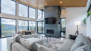 A modern living room with a large gray sofa, a flat-screen TV mounted on a gray wall, and a fireplace. - Spa, Views & Nature | Panoraama Finlandais (Saint-Côme)
