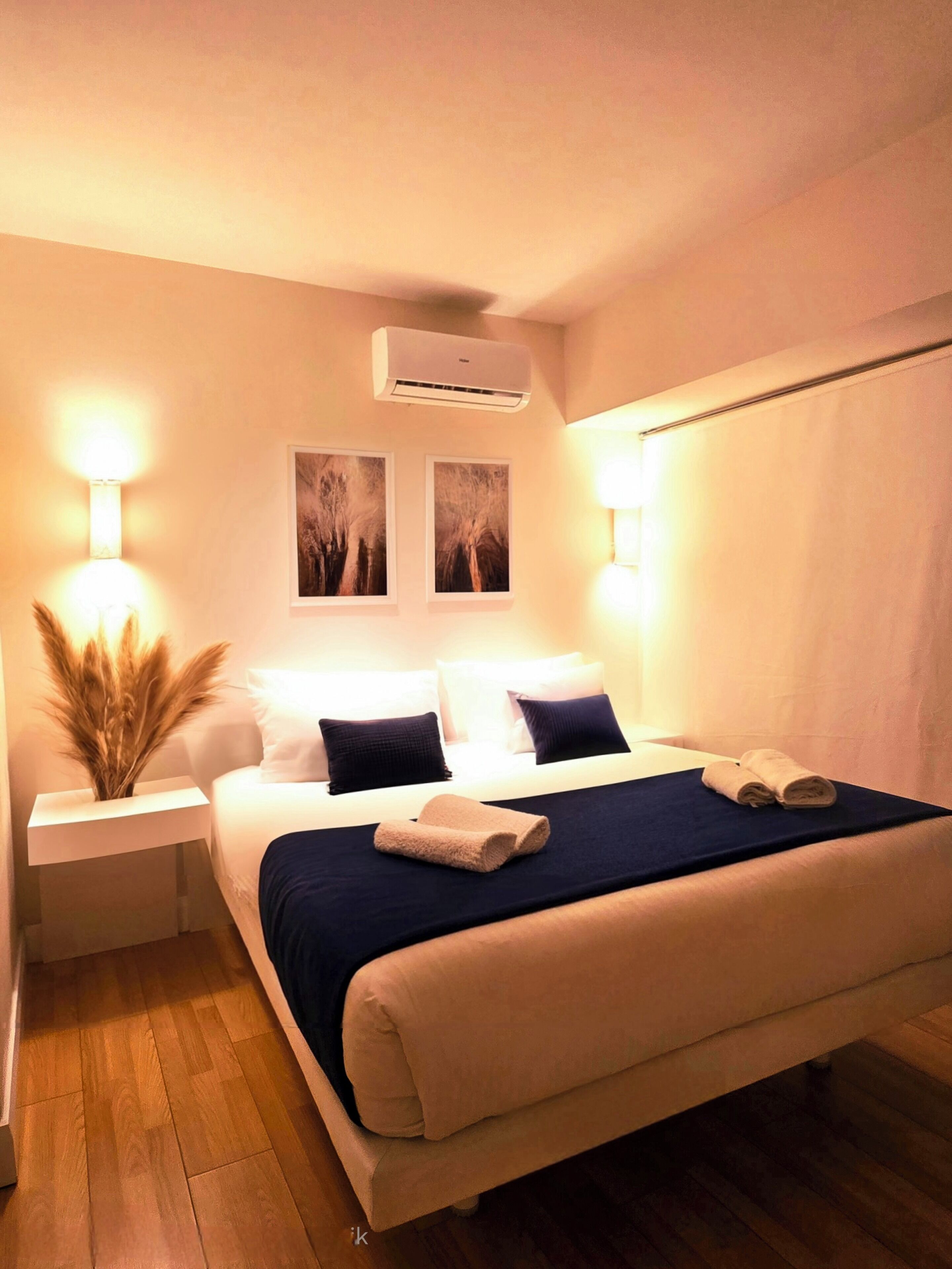 Una cama bien tendida con una colcha azul oscuro y dos almohadas, una lámpara de noche y un aire acondicionado instalado en la pared.
