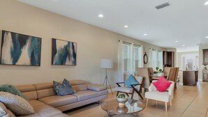 A spacious living room with a beige sofa, a coffee table, and a dining area with a white chair and a red pillow. - 25% Off 8BR Villa! Pool/Spa.  Game & Movie Rooms (Davenport)