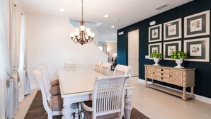 Un coin repas avec une table et des chaises blanches, un lustre et un mur d’accent bleu foncé orné de tableaux encadrés.