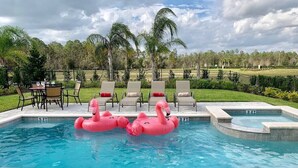 Une piscine avec deux bouées en forme de flamants roses, un espace détente avec des chaises et une table, et une vue sur un champ verdoyant parsemé d’arbres.