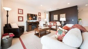 Un salon comprenant un canapé, un fauteuil, une table basse et une cheminée. Des tableaux encadrés ornent le mur et une lampe est posée sur le sol.