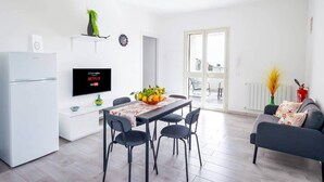 Una cocina moderna con refrigerador empotrado, un área de comedor con mesa y sillas, un televisor de pantalla plana mostrando servicios de streaming y vista al exterior a través de una puerta de cristal.