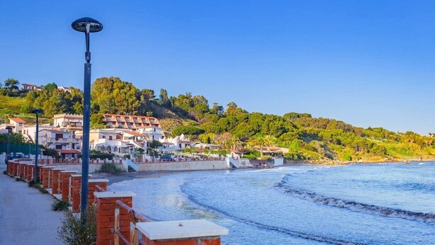 Un pueblo costero con edificios en una ladera, una playa y una calle con barreras de ladrillo rojo.