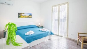 Un dormitorio con cama, mesita de noche, silla, ventana con cortinas y un cuadro en la pared.
