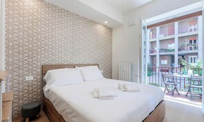 Una camera d'albergo con un letto, uno sgabello, un termosifone e un balcone con tavolo e sedie.