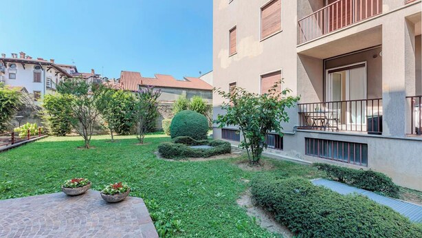 Ein Innenhof mit Pflasterfläche, ein kleiner Garten mit einem ordentlich geschnittenen Busch und ein Gebäude mit Balkonen und Fenstern.