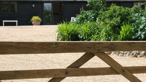 A wooden fence in front of a building with a gravel driveway and a garden area with various plants. - Solo Country Getaway in Kent (Ashford)