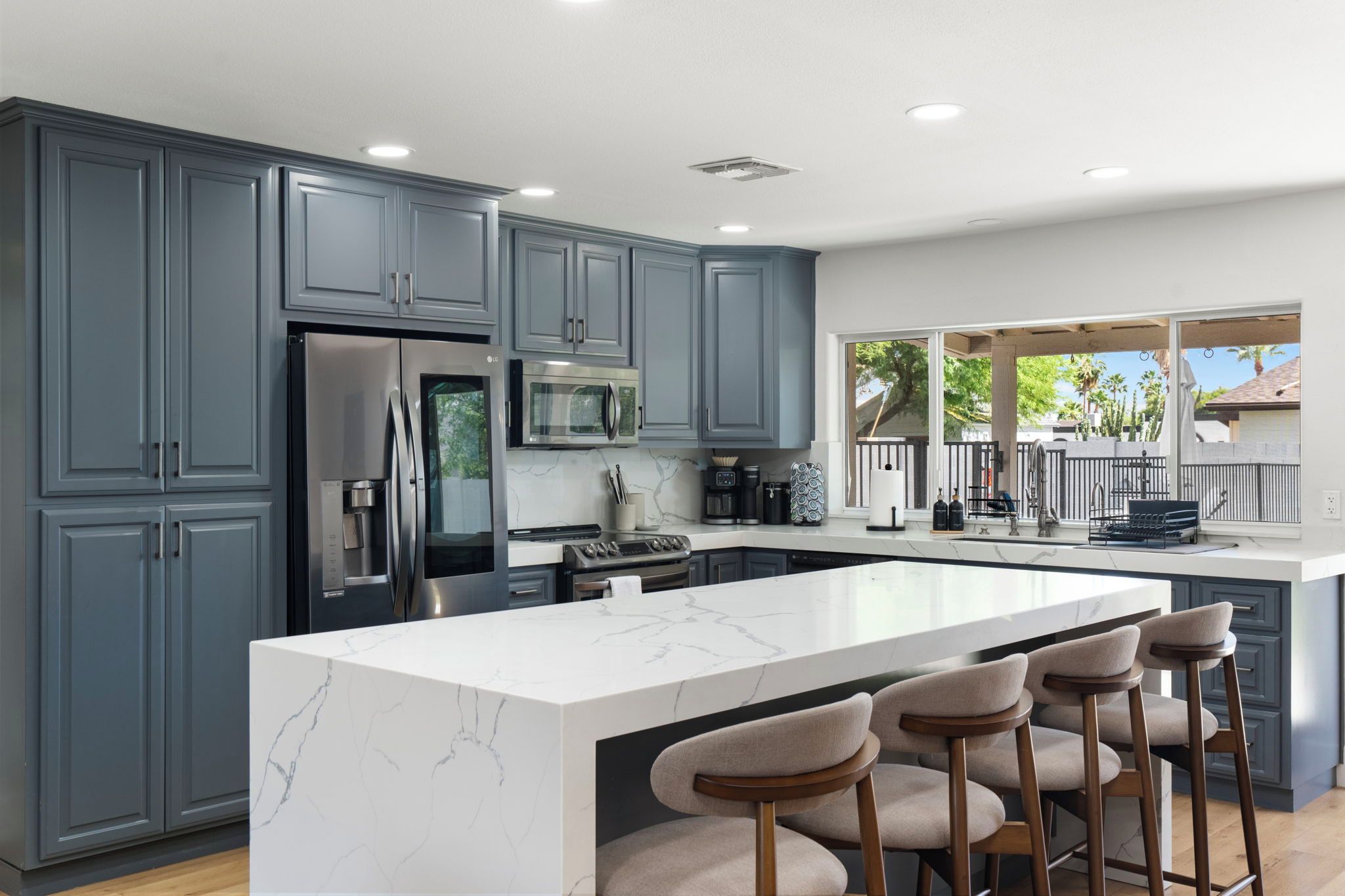Una cocina moderna con armarios grises, una isla grande con taburetes de bar, electrodomésticos de acero inoxidable y vistas al exterior.