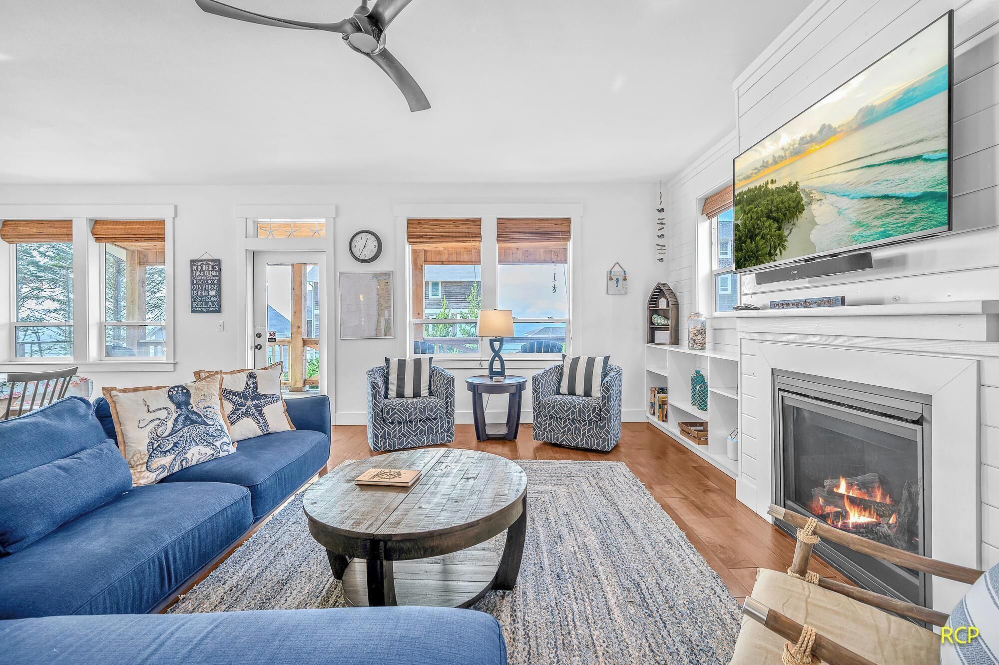 Un salon avec un canapé bleu, des fauteuils à motifs, une table basse ronde, une cheminée et un téléviseur fixé au-dessus.